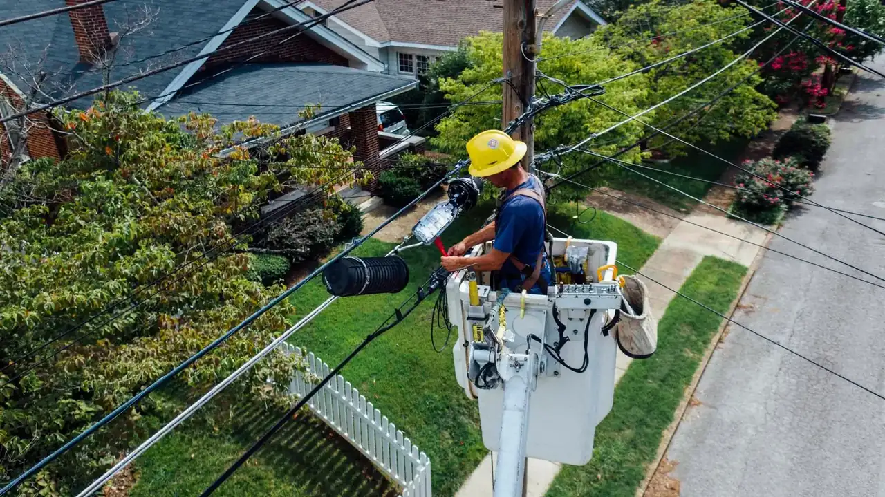 Worker in a hard hat riding an aerial lift bucket high above a tree-lined suburban street