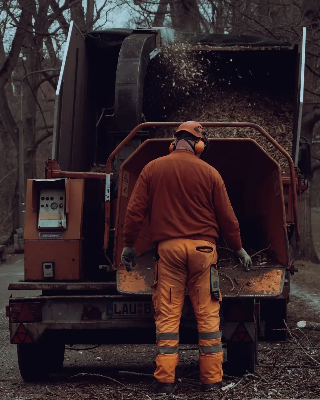 Worker in hi-vis pants feeding brush into the hopper of a truck-mounted chipper as chips blow into the box