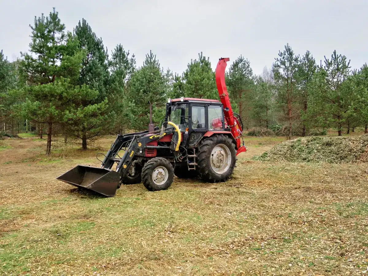 Tractor-mounted wood chipper working beside a fresh pile of chips at a wood lot